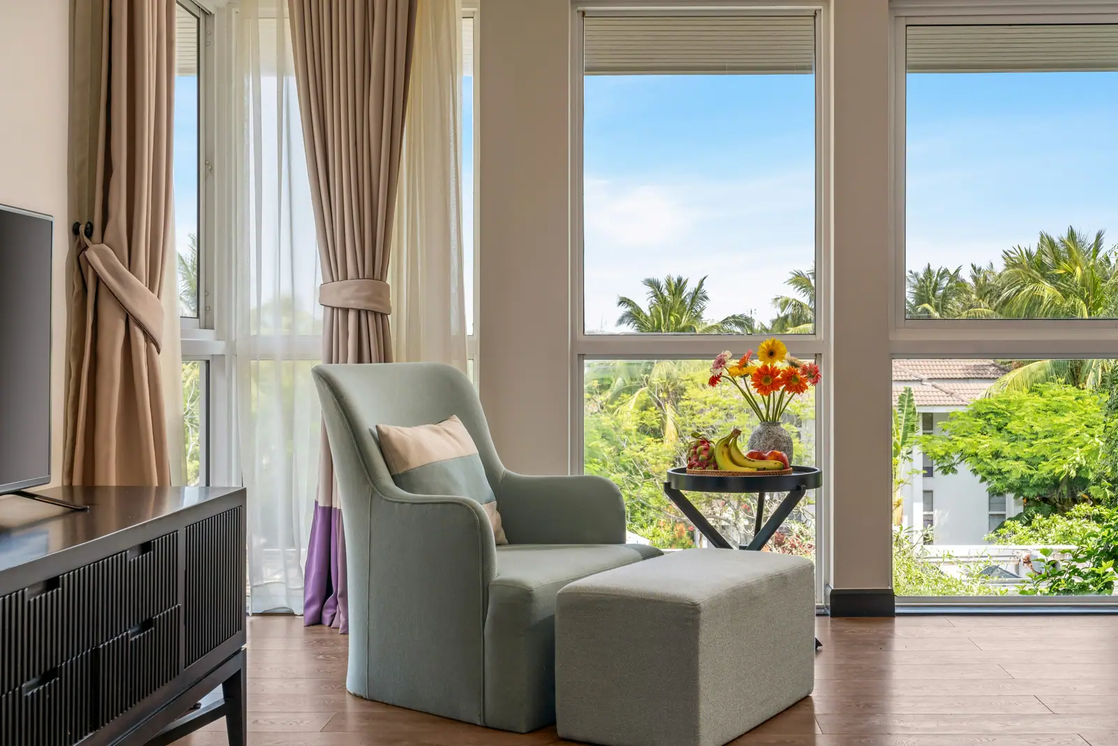 A sage armchair and ottoman at a corner of tall windows looking onto palms
