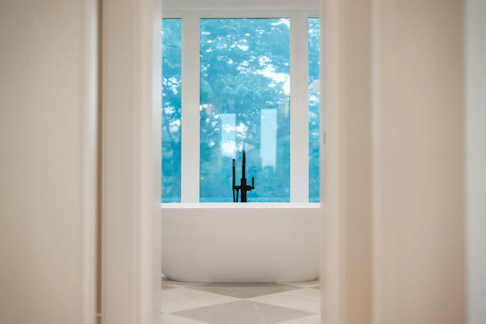 A freestanding white tub under a tall window full of green trees