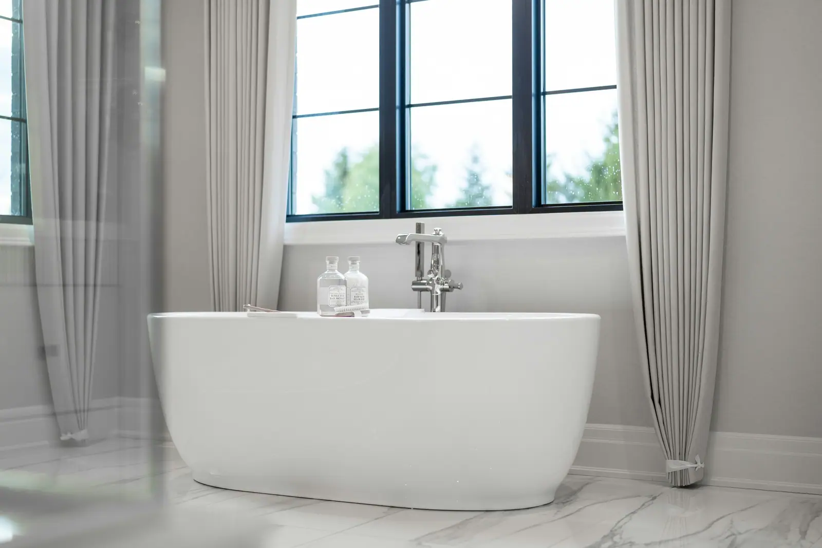A freestanding white tub on marble beneath a tall window with sheer curtains